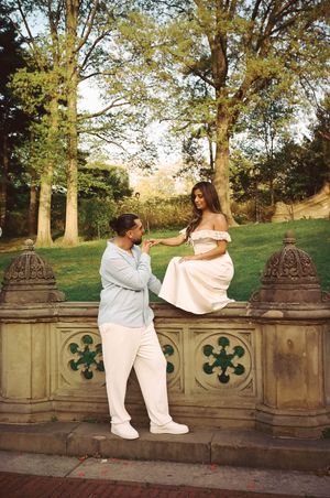 Romantic couple at Central Park stone terrace, woman in white dress sitting on wall while man gazes up at her.