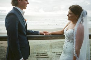 Bride and groom share a tender moment by the ocean, bride in lace gown with veil, groom in navy suit.