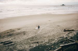 Aerial view of a couple in wedding attire walking on a vast sandy beach with ocean waves in the background.