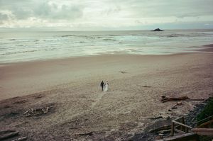 A lone person walks on a vast empty beach with calm ocean waves and overcast skies.