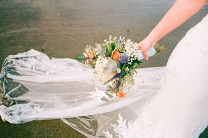 Bride holding a colorful wildflower bouquet with wedding veil spread across a dark sandy beach.