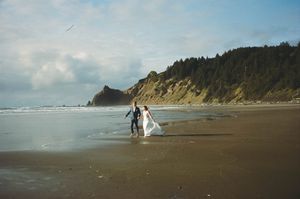Bride and groom walking hand in hand along a scenic Pacific Northwest beach with forested cliffs.