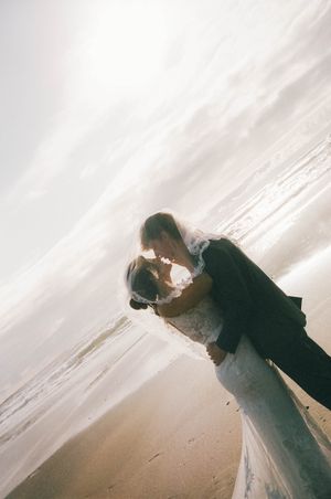 Bride and groom share a romantic kiss on a sunlit beach, with the bride's veil flowing in the breeze.