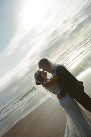 Bride and groom sharing a romantic kiss on a sunlit beach, with ocean waves and a glowing sky in the background.