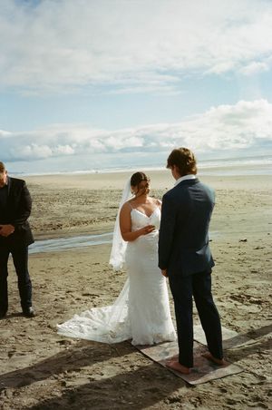 Beach wedding ceremony with bride in lace gown and veil exchanging vows with groom in navy suit.