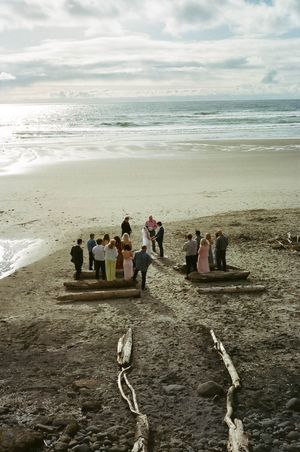 Small wedding ceremony on a scenic beach with driftwood logs, ocean waves, and dramatic cloudy sky backdrop.