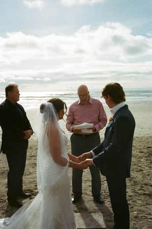 Bride and groom exchange vows in a beach wedding ceremony officiated by a man in a pink shirt.