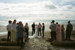 Beach wedding ceremony with small group of guests gathered around couple at shoreline under cloudy sky.