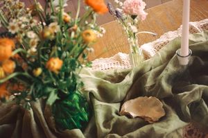Rustic wedding table decor featuring orange and pink flowers in a green vase on sage green linen fabric.