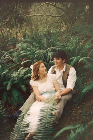 Couple sitting together among lush green ferns in a forest, woman in white dress, man in vest smiling at each other.