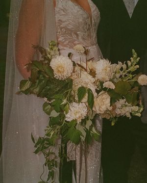 Close-up of a white lace wedding dress with an elegant bouquet of white dahlias and greenery.