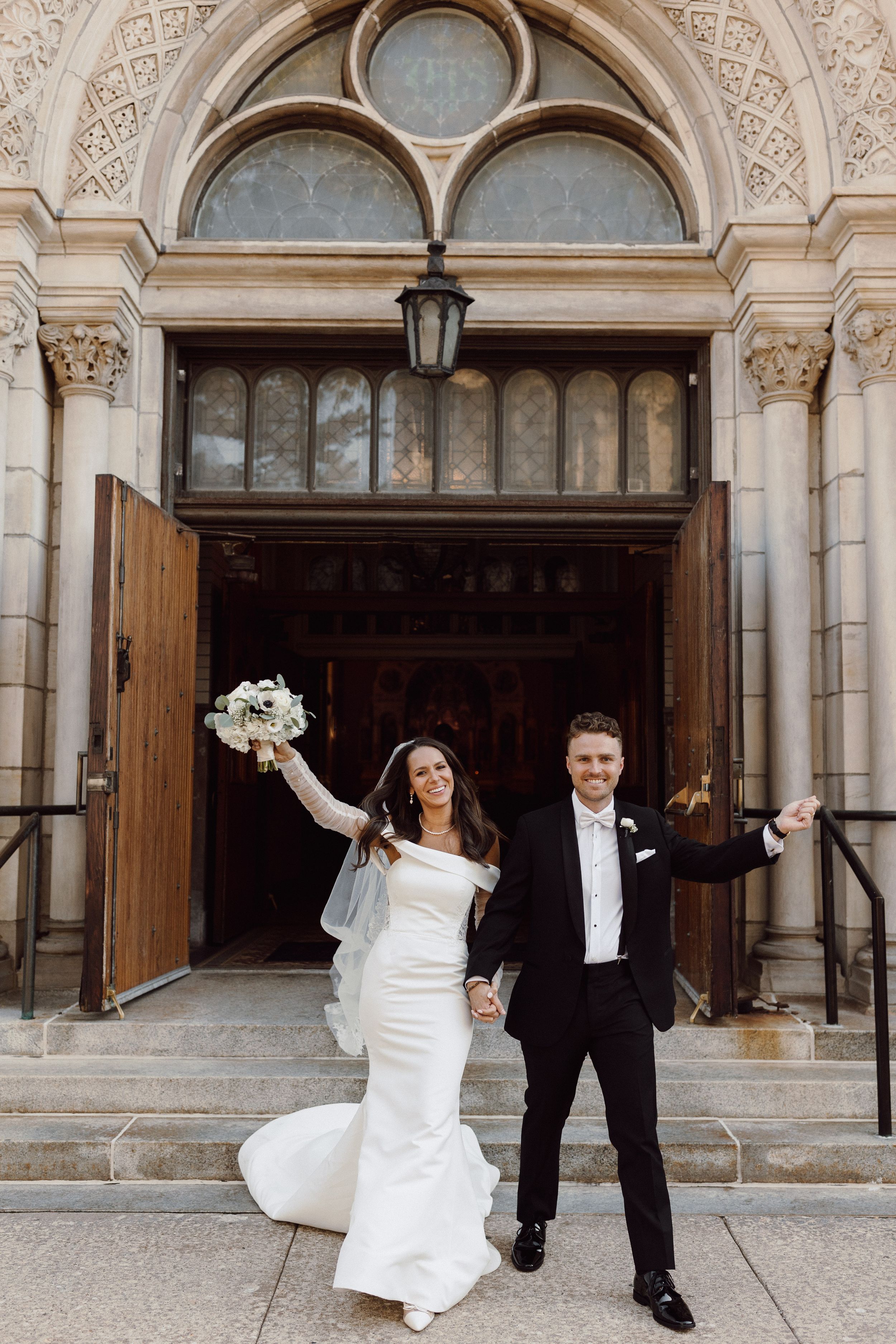 Newlyweds celebrate outside ornate stone church with dramatic architectural details.