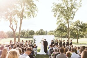 Outdoor wedding ceremony under trees with guests seated in white chairs at sunset.