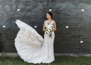 Elegant lace wedding dress flows against dark brick wall background.