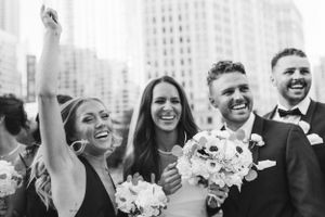 Joyful wedding party celebrating with raised arms against a bright city backdrop.