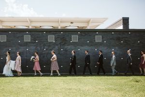 Wedding party walks in line against a modern dark wall on a sunny day.