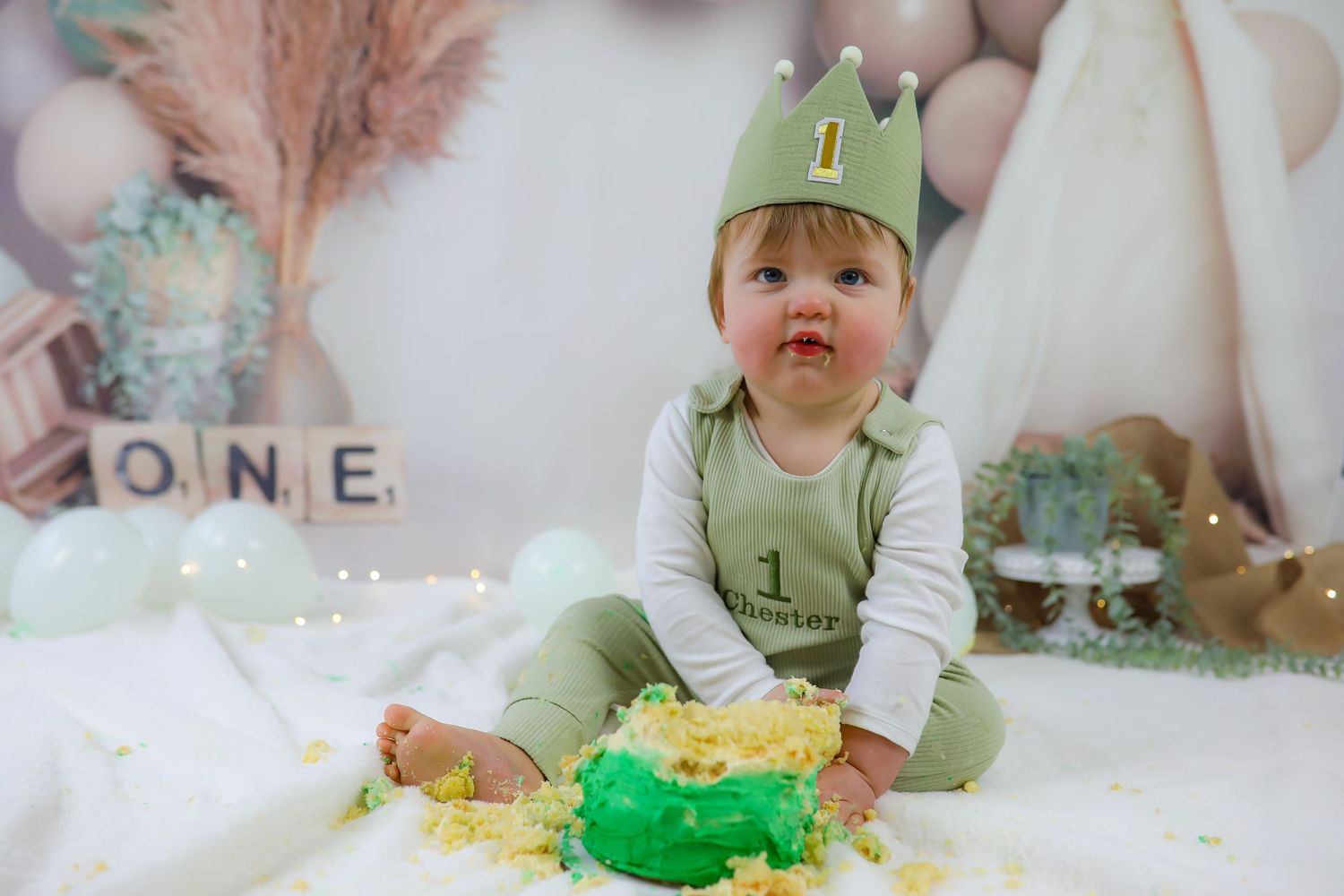 A one-year-old in a green crown and overalls sits on white fabric near a smashed green cake and letter blocks.