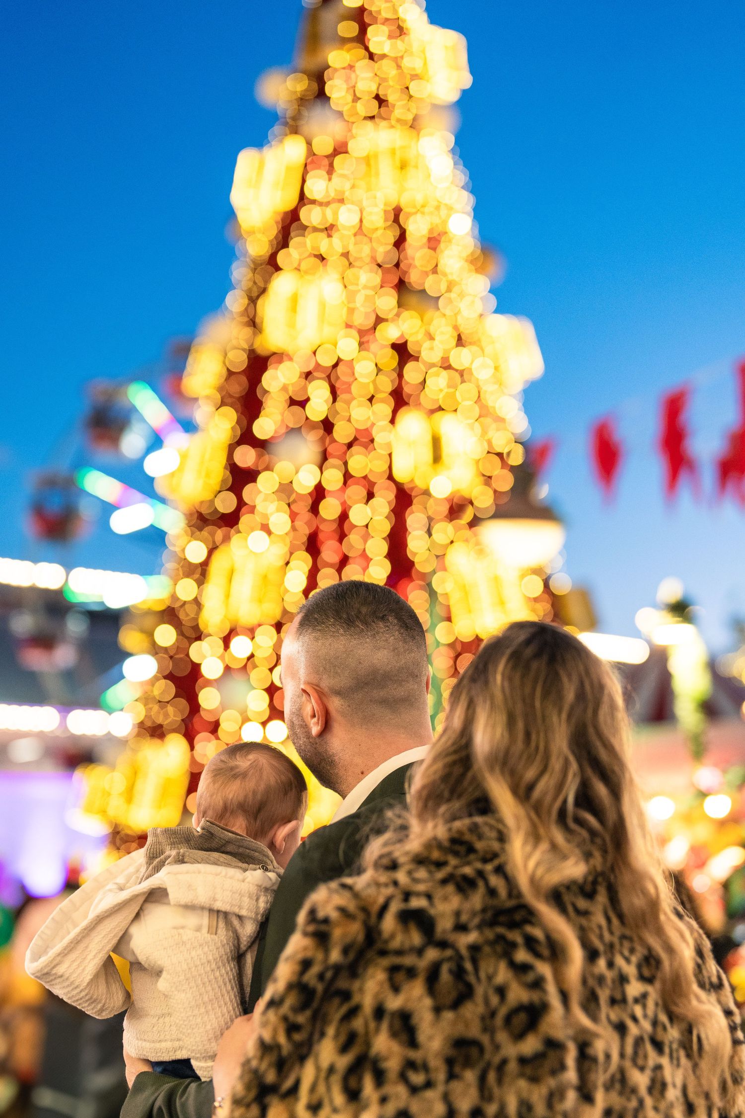 Family gazes at a glowing Christmas tree decorated with warm bokeh lights against a deep blue evening sky at a festive fair. Βάπτιση Αθήνα