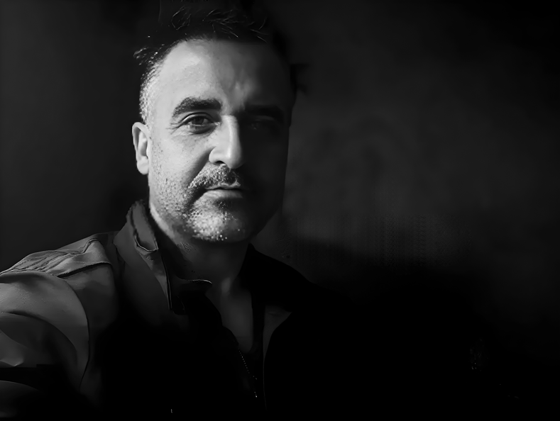 Dramatic black and white portrait of a person with facial hair and a contemplative expression in low-key lighting.