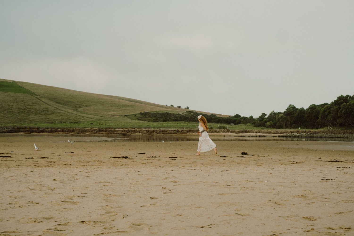 Distant silhouette in white dress stands alone on vast empty beach with green hills in background.