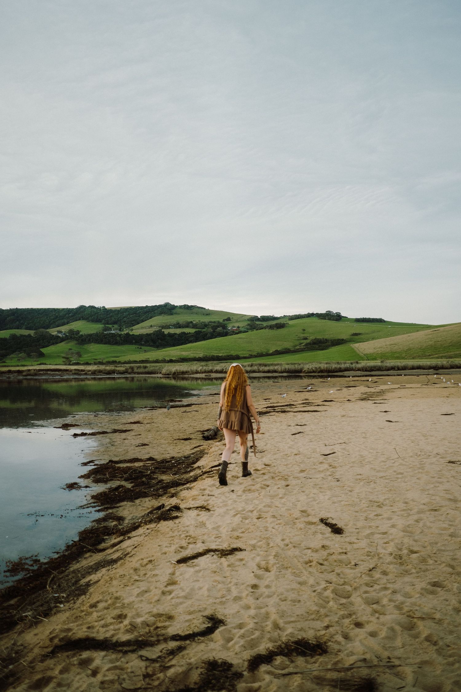 Solitary figure walks along shoreline with rolling green hills in the misty background.