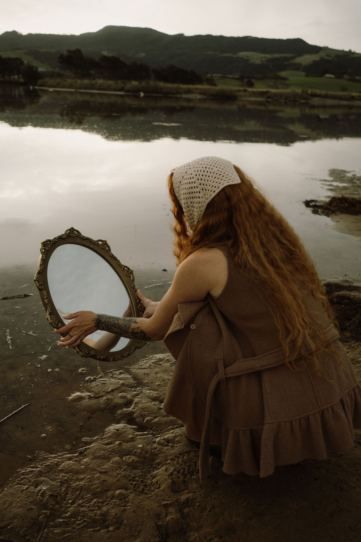 Figure in brown dress holds vintage mirror by waterside with hills reflected in calm water.