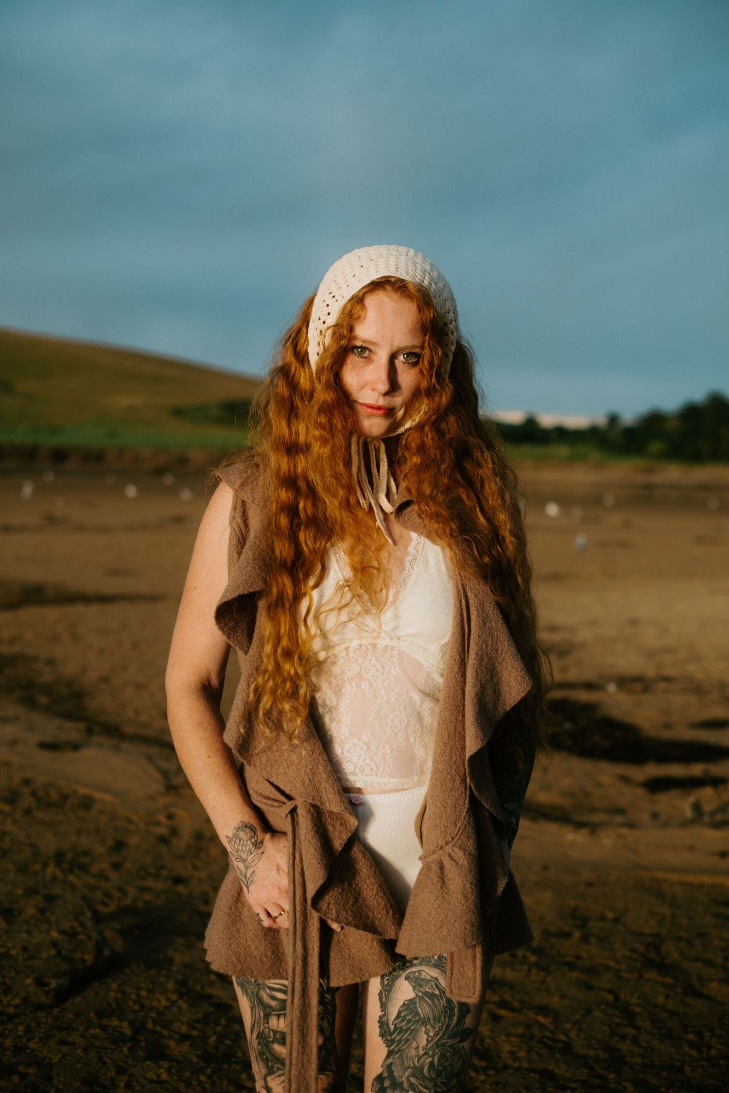 Person in knit beanie and brown cardigan stands on beach during golden hour.