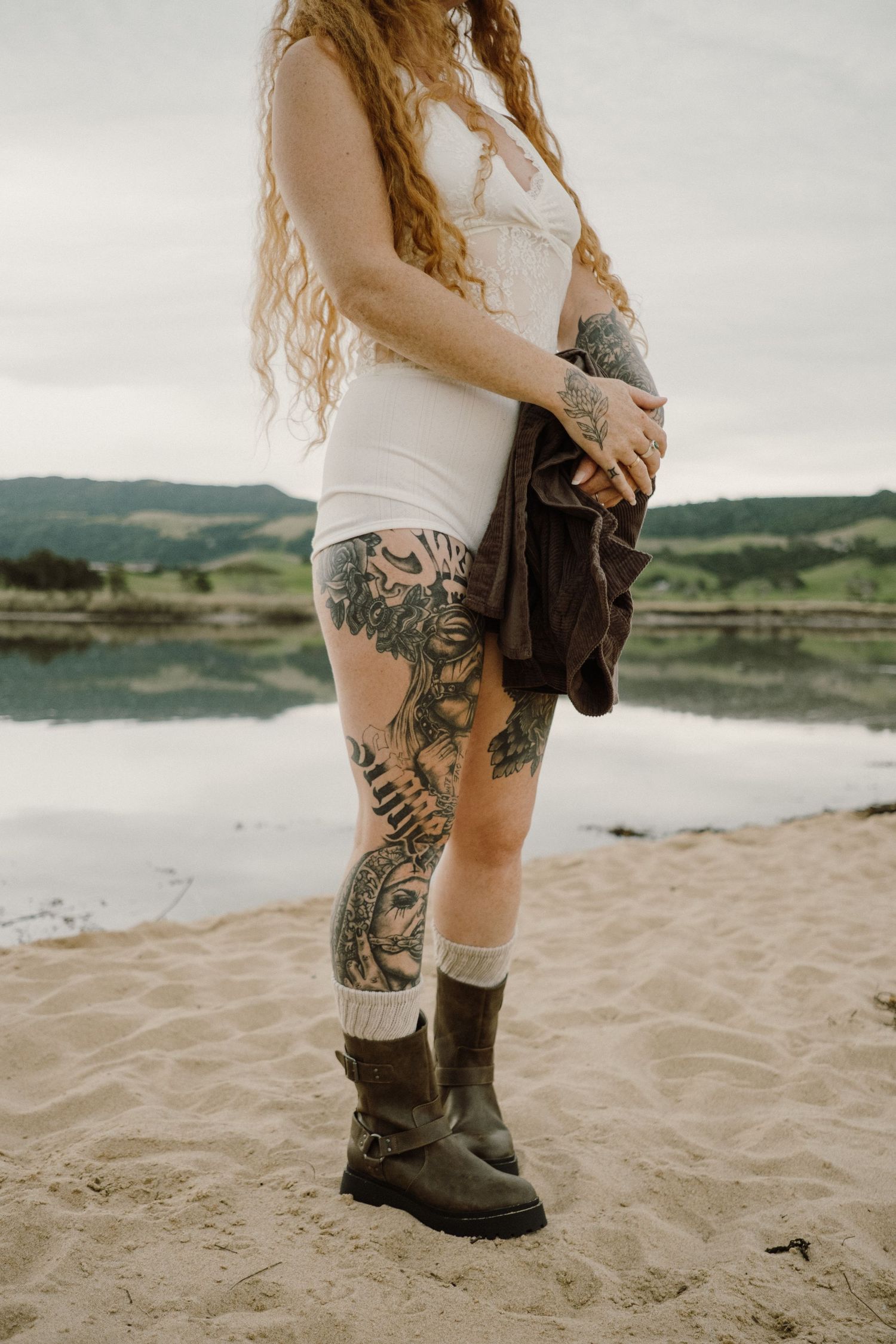 Close-up of leg tattoos and combat boots on sandy beach with water and hills in background.
