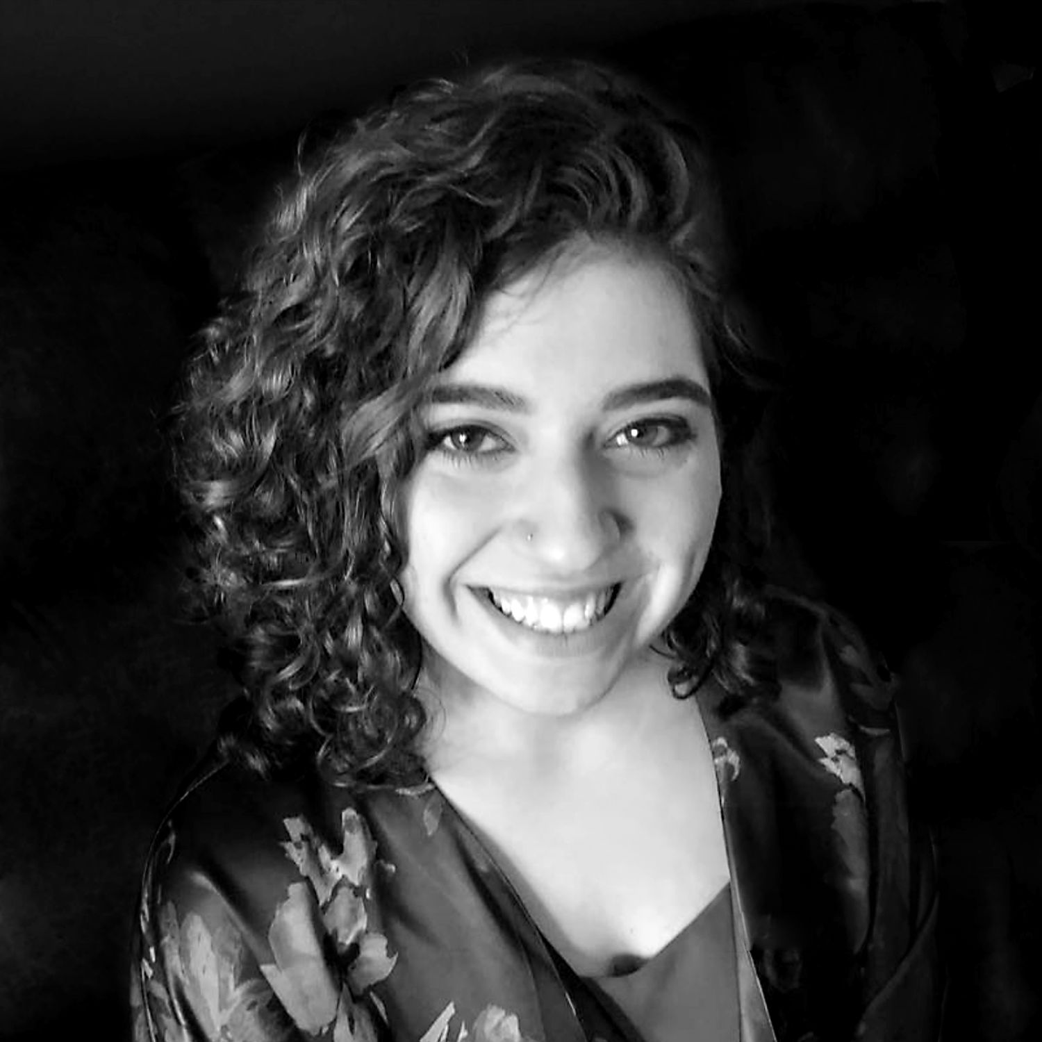 Black and white portrait of a smiling person with dark curly hair wearing a patterned top against a dark background.