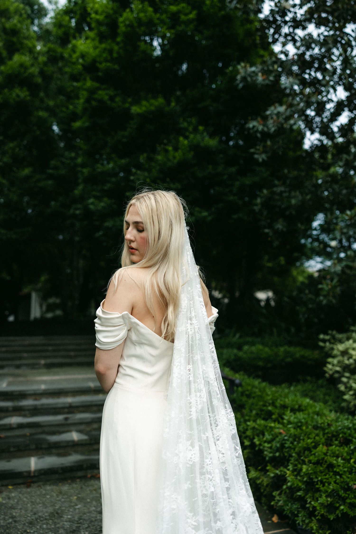 Bride in off-shoulder dress with long veil poses in garden setting.