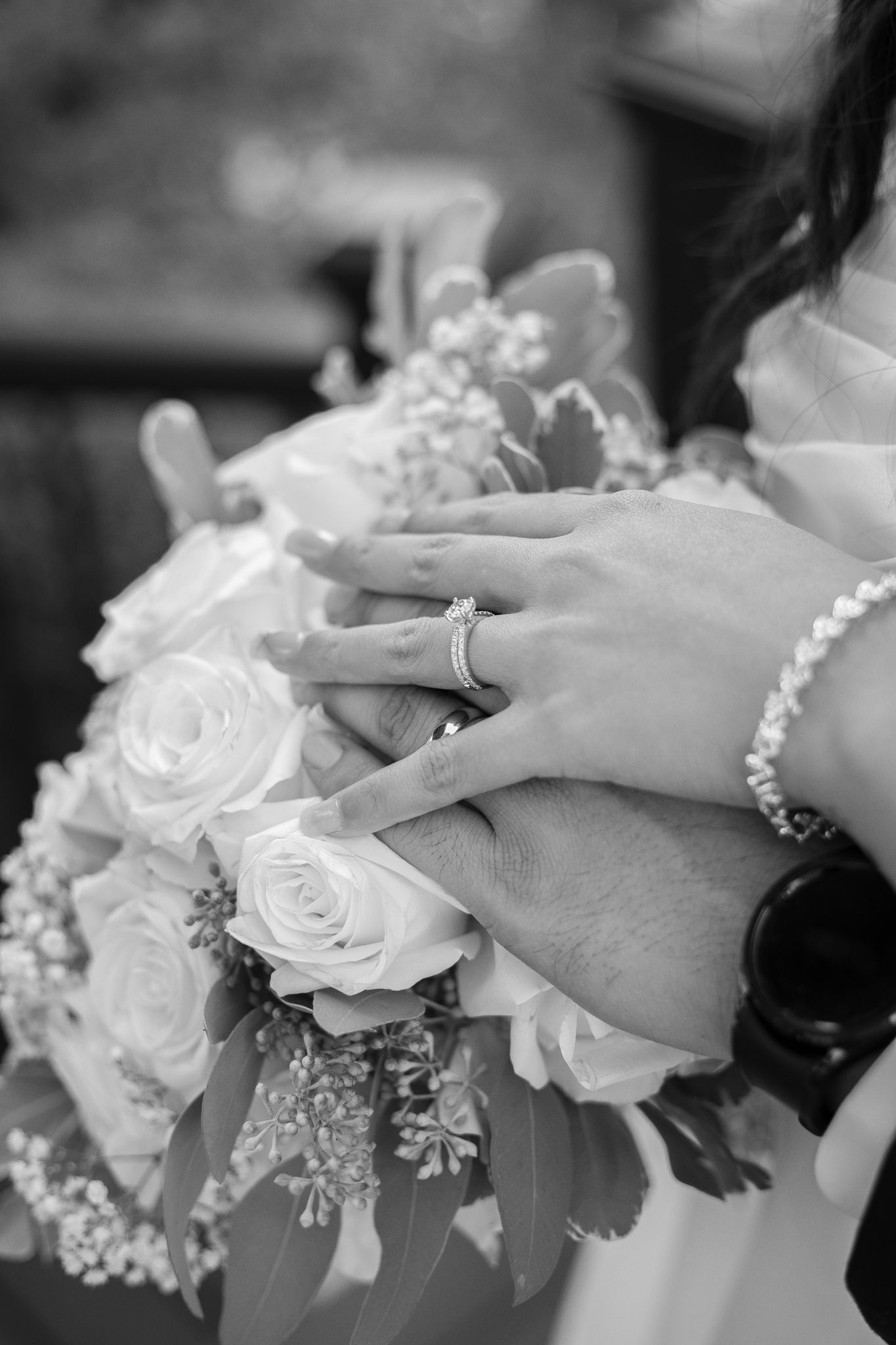 A black and white close-up of wedding rings and hands holding a rose bouquet.