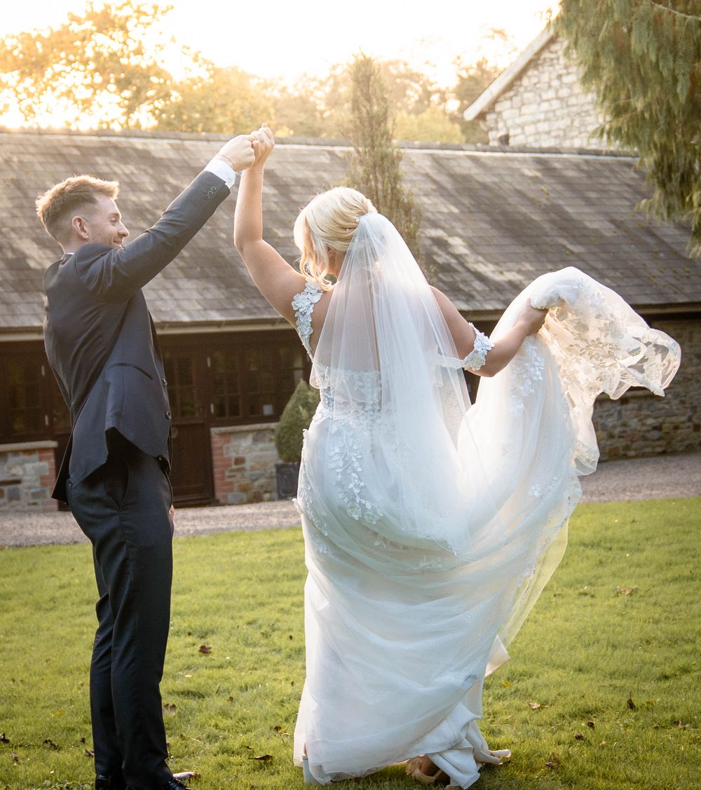 Joyful wedding couple dancing and twirling in front of a rustic building at sunset.