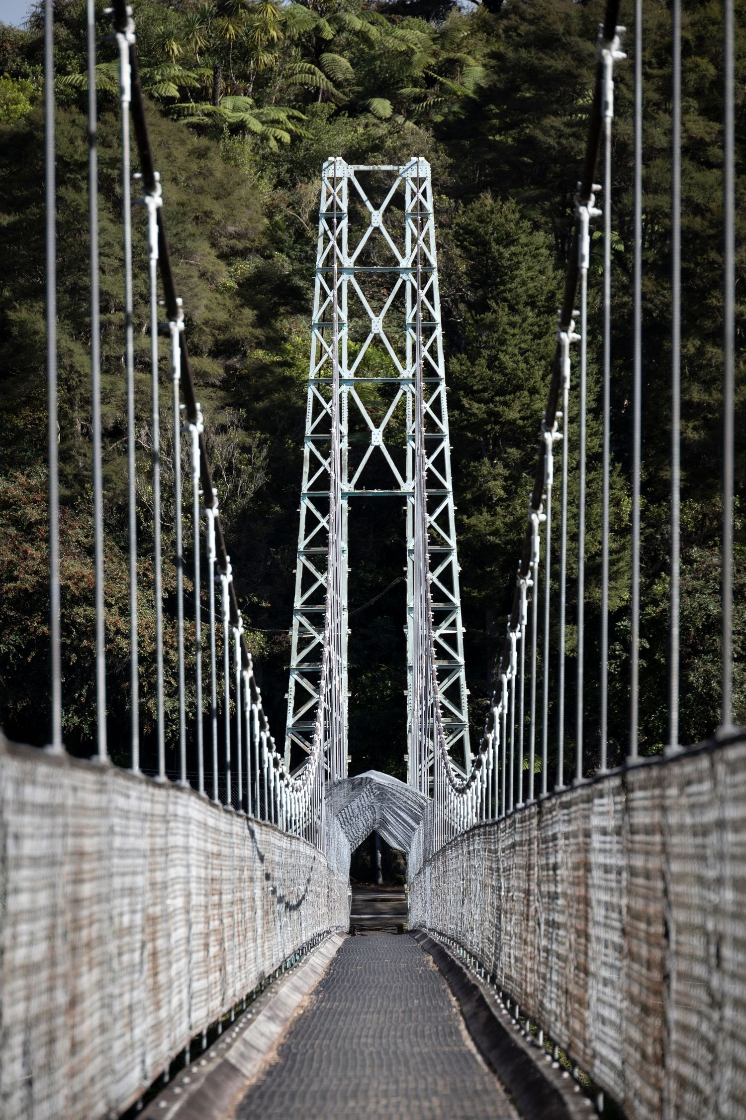 Suspension bridge with white lattice tower and cables, leading through forested hills on a bright day.
