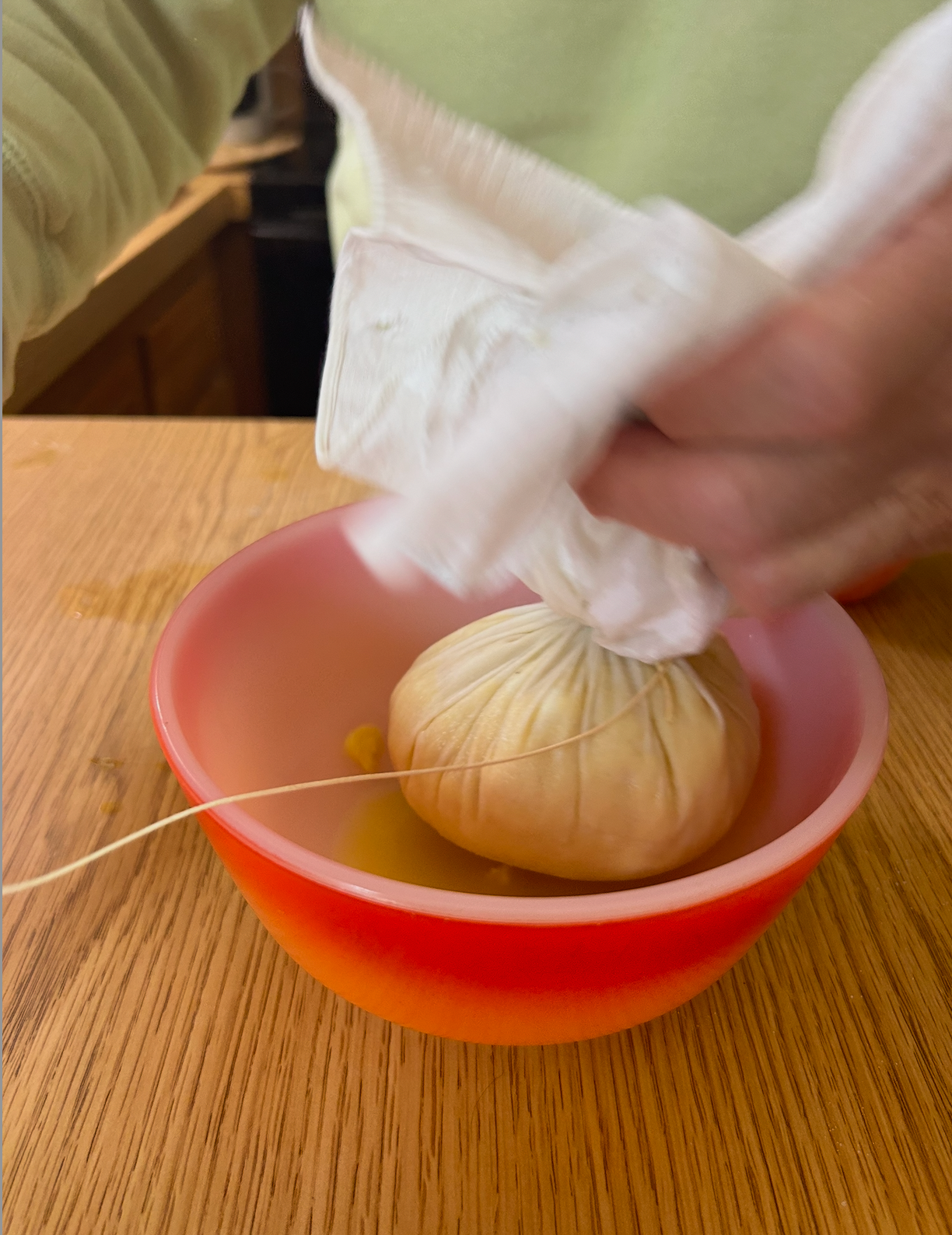 Hands squeeze a cheesecloth filled with strained ingredients over an orange and red bowl on a wooden table.