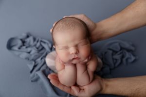 Sweet newborn baby sleeps peacefully in supportive hands on gray textile backdrop during professional photo session.