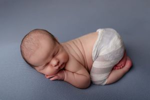 Peacefully sleeping newborn baby wearing a white diaper rests on blue textured backdrop in fetal position.