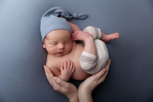 Sleeping newborn baby wearing a soft gray hat being gently cradled in hands against gray backdrop.