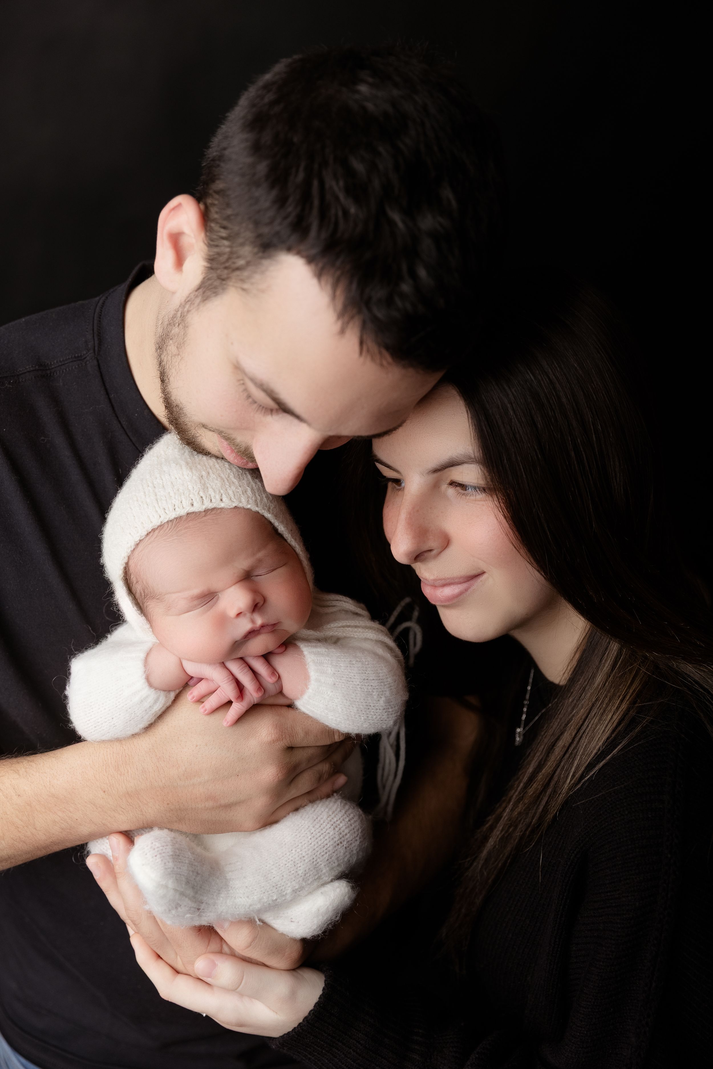 A tender family portrait shows parents gazing lovingly at their newborn baby wrapped in a white knit blanket.