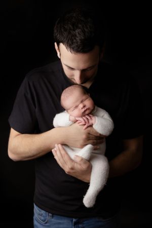 A tender moment captured in low key lighting shows a parent in a black shirt holding a sleeping newborn in white clothing.