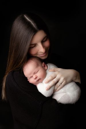 Tender portrait of a mother in black holding her sleeping newborn baby against dark background.