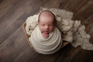 Sleeping newborn swaddled in white blankets nestled in wooden bowl on dark hardwood floor with lace fabric.