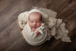 A sleeping newborn wrapped in cozy white knit blankets lies in a wooden basket with a small plush toy companion.