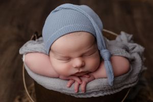 Peaceful sleeping newborn in gray knit bonnet lying in a wooden basket with soft gray fabric.