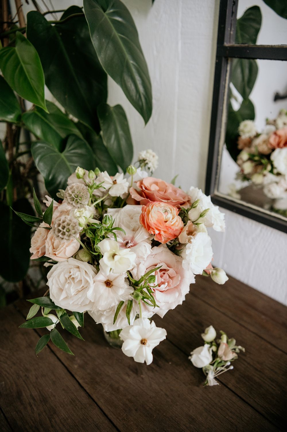 Elegant bridal bouquet with pink and white roses sits on dark wooden surface next to window with tropical plants.