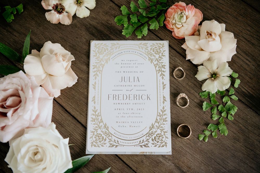 Elegant wedding invitation with gold foil details surrounded by blush roses, wedding rings and greenery on wooden surface.