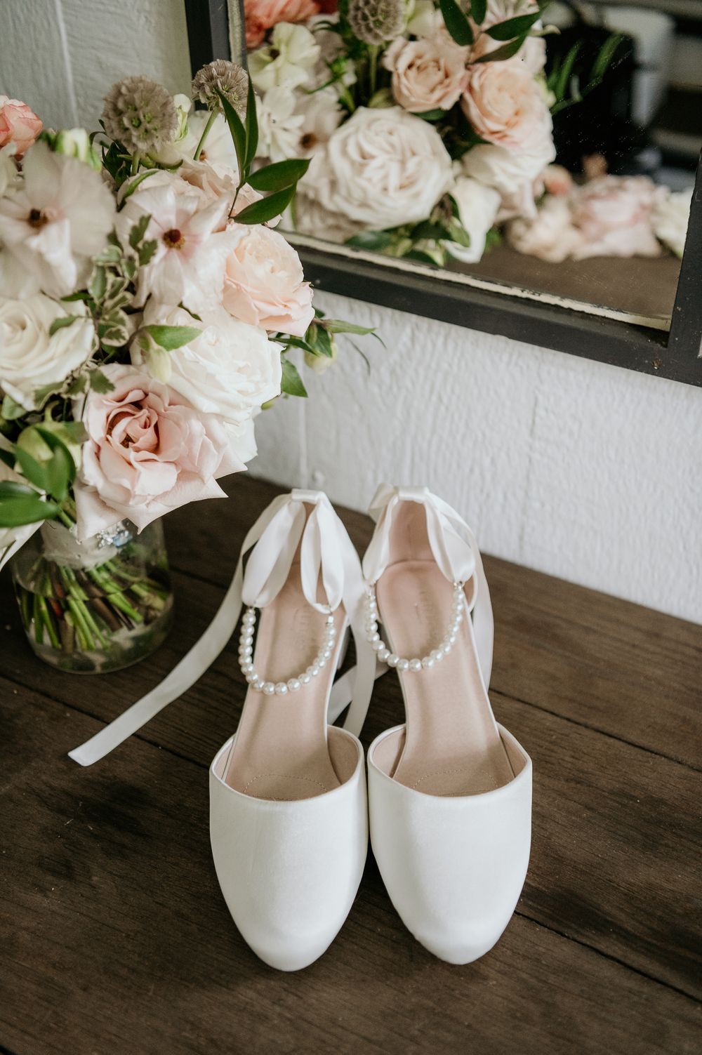 White bridal shoes with ankle straps displayed next to a soft pink rose floral arrangement on a dark wooden surface.