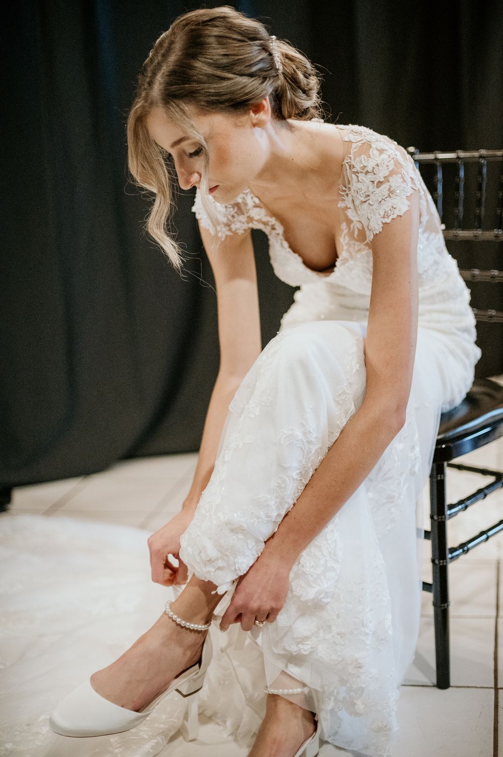 A bride in a lace wedding dress with sheer sleeves adjusts her white shoe while sitting on a dark chair.