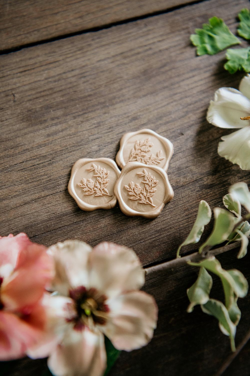 Delicate wax seals with floral designs on wooden surface surrounded by pink and white flowers and greenery.