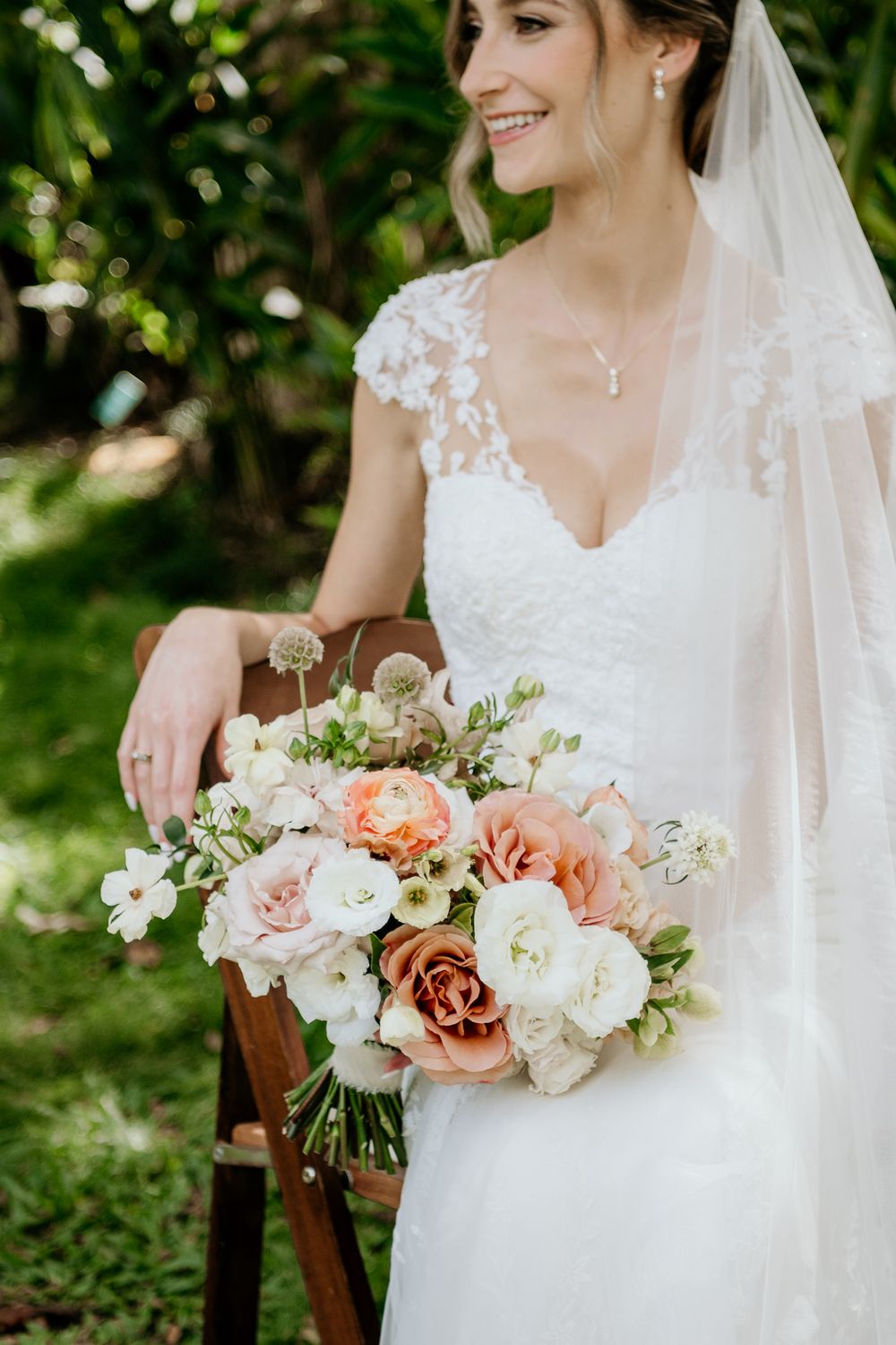 A bride in a lace cap sleeve wedding dress holds a beautiful bouquet of peach and white roses in a garden setting.