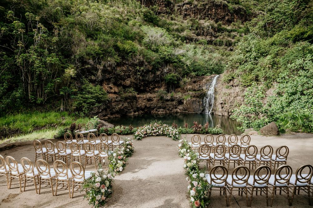 White garden chairs arranged for an outdoor wedding ceremony with a scenic waterfall backdrop in a lush tropical setting.